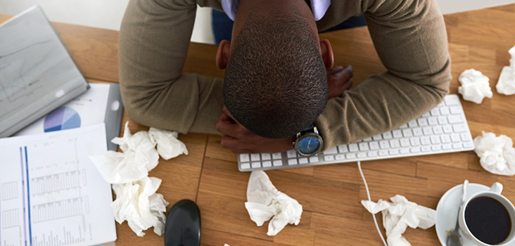 Depressed man with his head on his keyboard and balled up tissue paper.