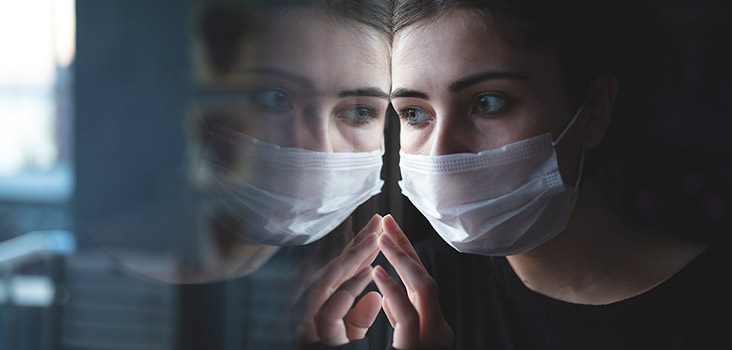 Woman wearing medical mask looks at her reflection through a glass window