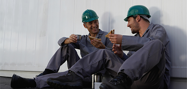 2 workers wearing green hard hats eating lunch and talking